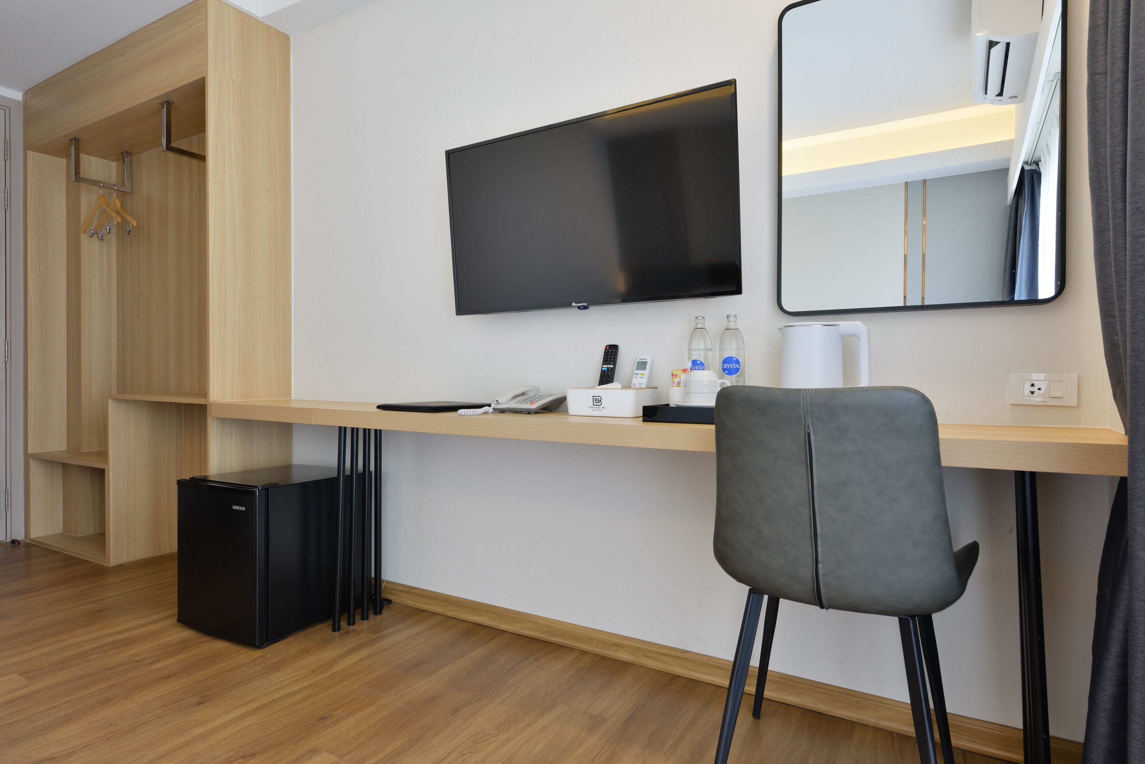 A hotel room with a wooden desk, a flat-screen TV, a mirror, a chair, a mini fridge, and a hanging coat rack.