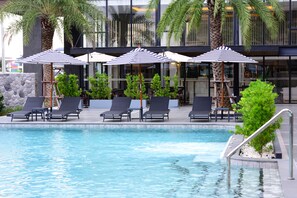 A pool area with lounge chairs and umbrellas, surrounded by palm trees and a modern building facade. - Grand BS Airport Hotel Suvarnabhumi (Bangkok)