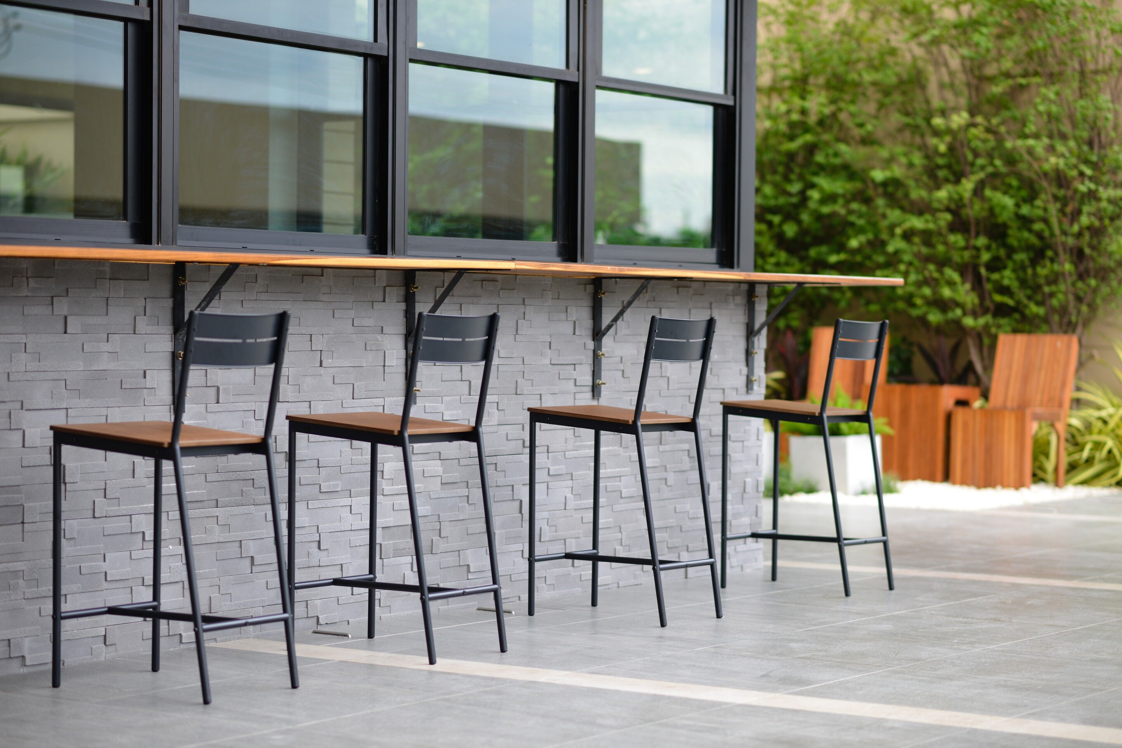 Un espacio moderno al aire libre con tres sillas y una mesa de madera frente a un muro de ladrillo, con una ventana arriba y vegetación en el fondo.