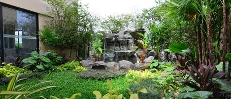 Un jardín con cascada de rocas, diversas plantas y un edificio con ventanas amplias.