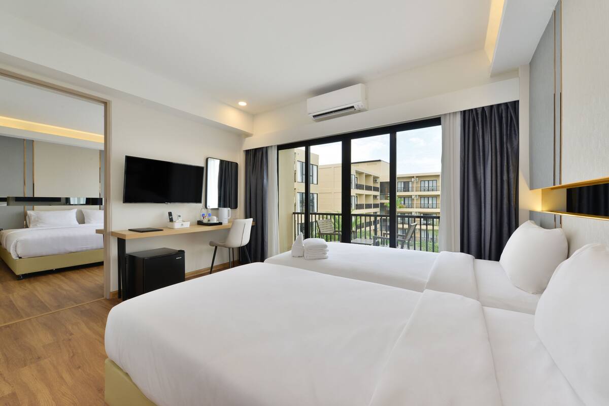 Ein Hotelzimmer mit zwei Betten, einem Schreibtisch, einem Stuhl, einem Flachbildfernseher und einem großen Fenster mit Blick auf Gebäude.