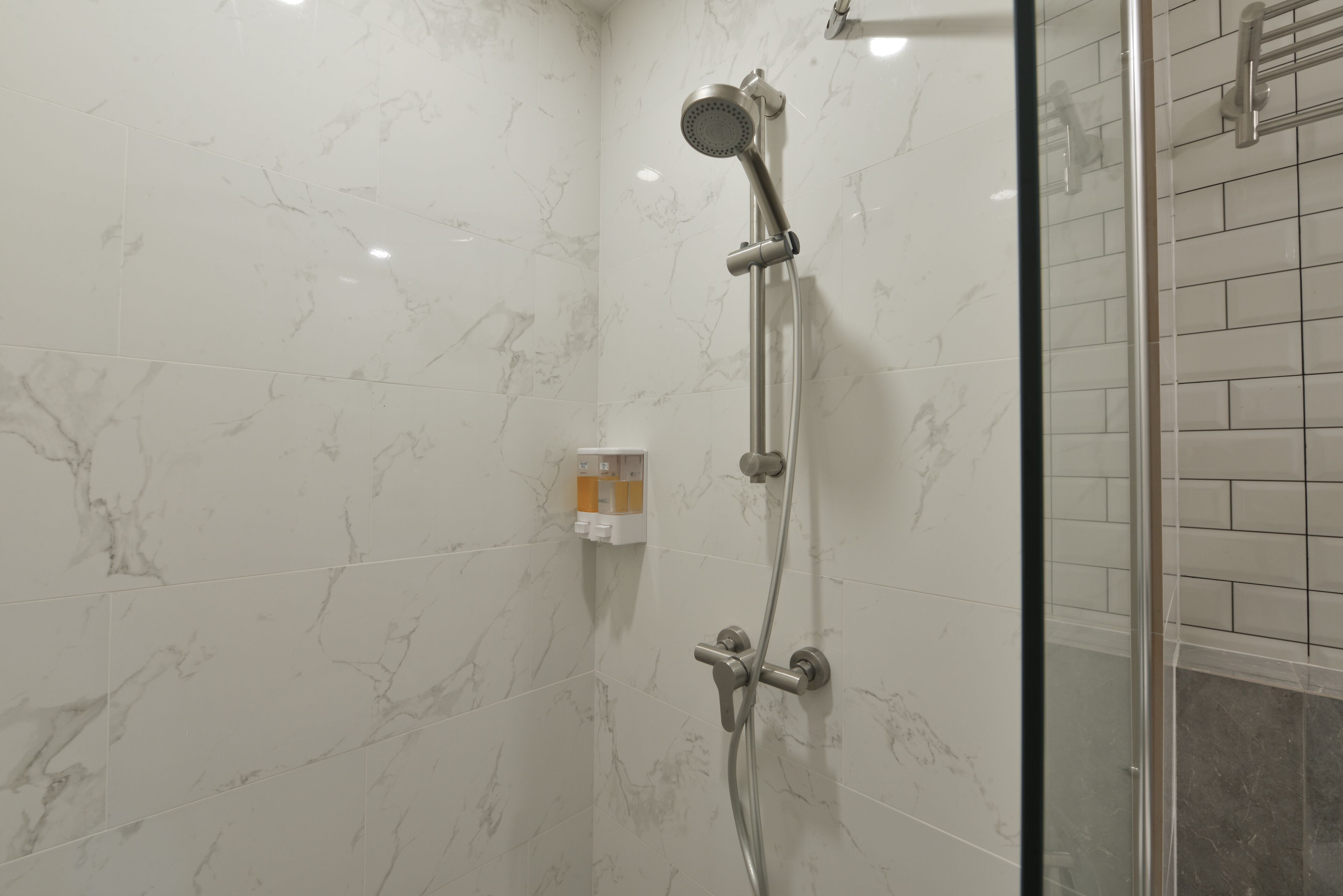 A shower with a handheld showerhead and a wall-mounted faucet, white marble tiles, and a glass shower enclosure.