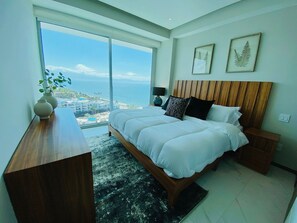 Un dormitorio con una cama grande, cabecera de madera, mesita de noche, escritorio y vistas al océano y edificios.