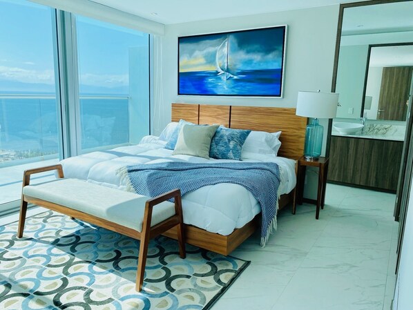 Un dormitorio moderno con una cama grande, cabecera de madera, vista al océano, un cuadro de un velero y un baño con encimera de mármol.
