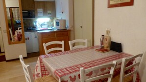 A dining area with a checkered tablecloth, white chairs, and a kitchen in the background. - Studio 2 cabins 4 pers. pets allowed, private parking (Valloire)
