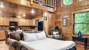 A cozy cabin interior with a large bed, wooden walls, a kitchen area, and a window with a view of greenery.