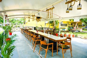 Een terras met een lange tafel en stoelen, omringd door potplanten en een geel gebouw op de achtergrond.