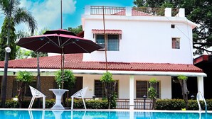A swimming pool with a white building in the background, a red umbrella, and two white chairs. - TGI Insignia Resort & Villa's (Bhopal)