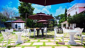 An outdoor patio with white metal chairs and tables, covered by red umbrellas, set on a patterned stone floor with green grass. - TGI Insignia Resort & Villa's (Bhopal)