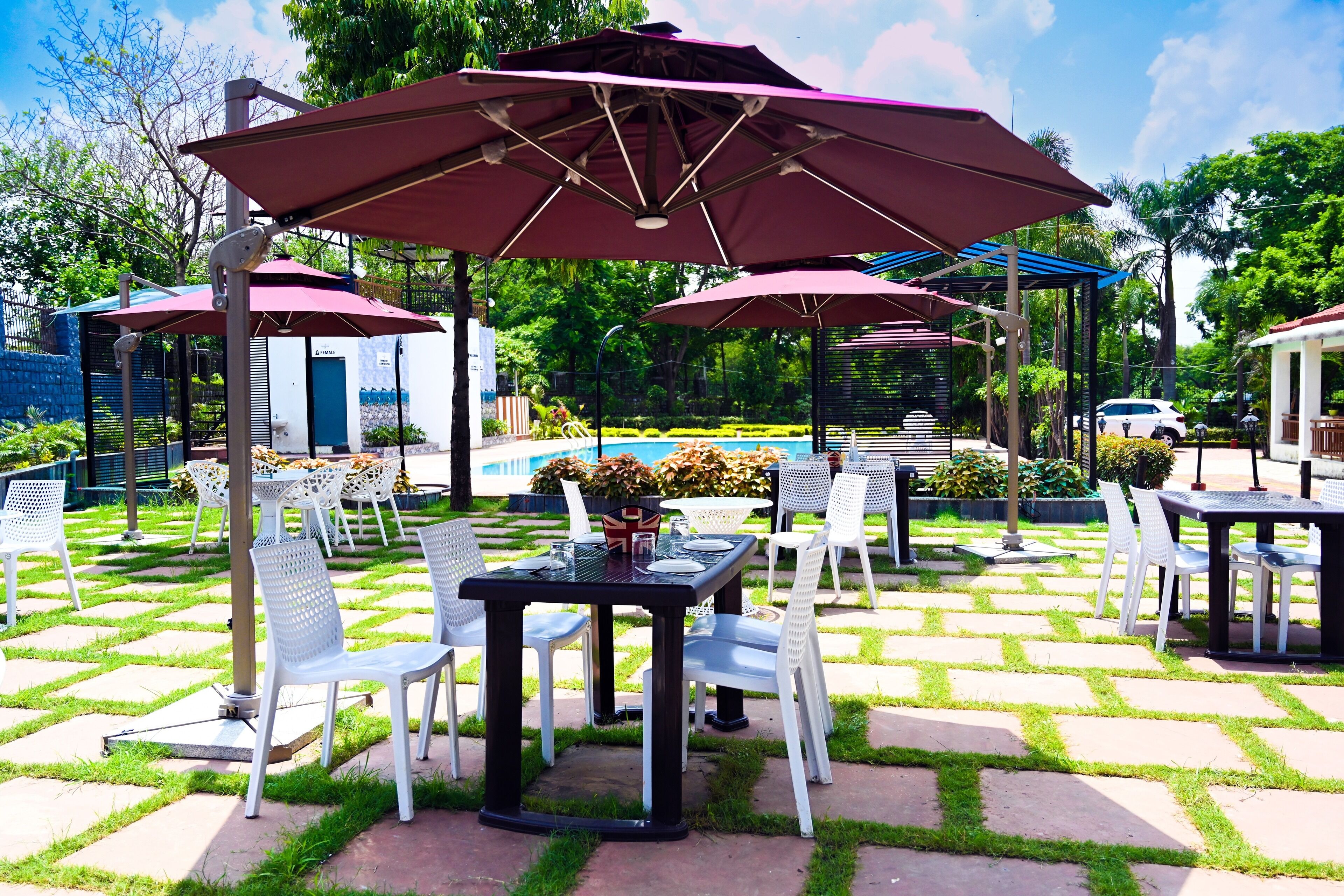 Un patio al aire libre con mesas y sillas bajo sombrillas, una piscina al fondo y un jardín.