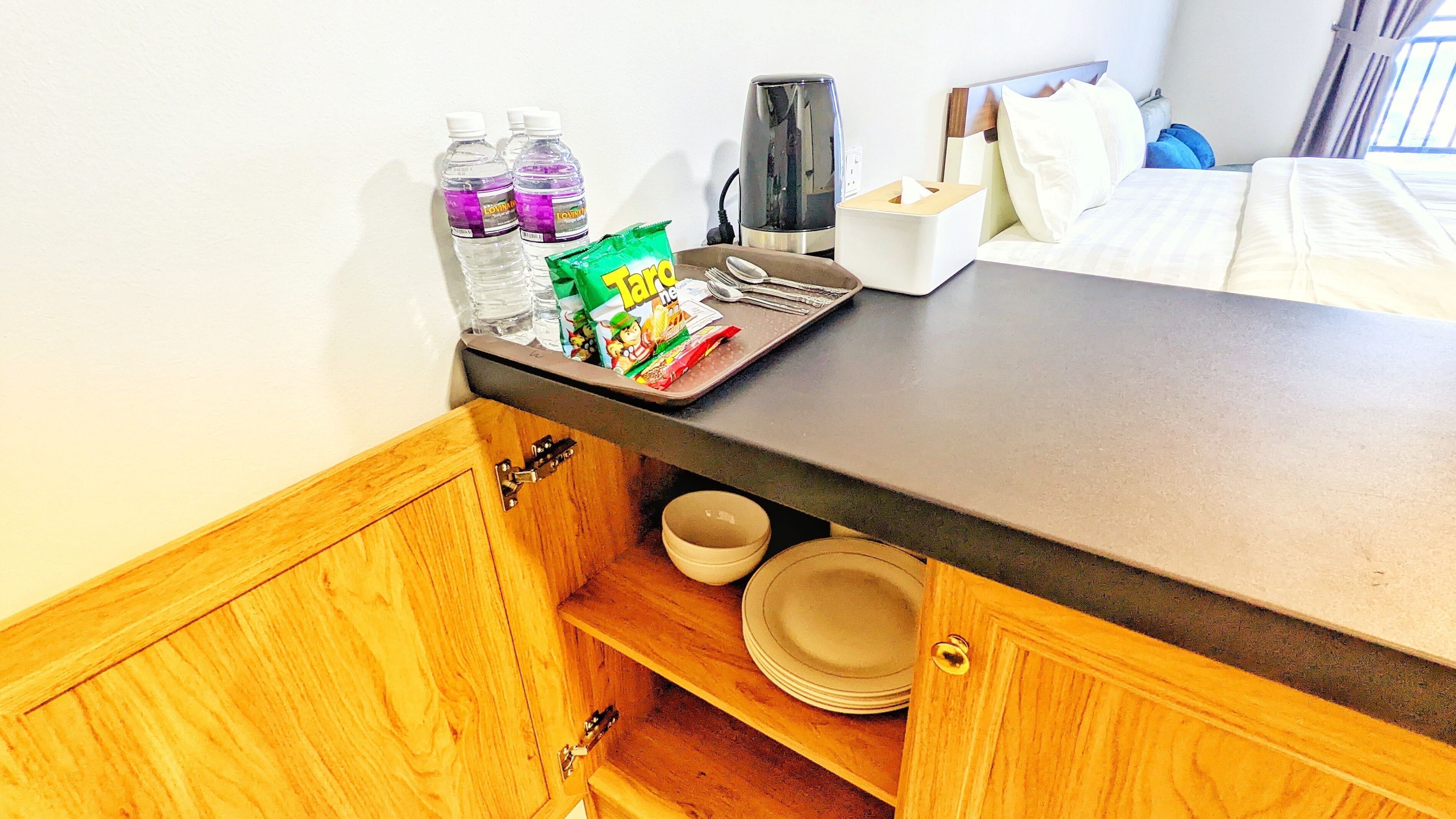 A hotel room with a wooden cabinet containing bowls and plates, a tray with water bottles, a coffee maker, and a snack package.