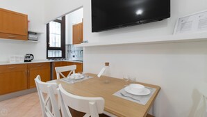 Une cuisine moderne avec des meubles en bois, un téléviseur noir fixé au mur et un coin repas comprenant une table en bois et des chaises blanches.