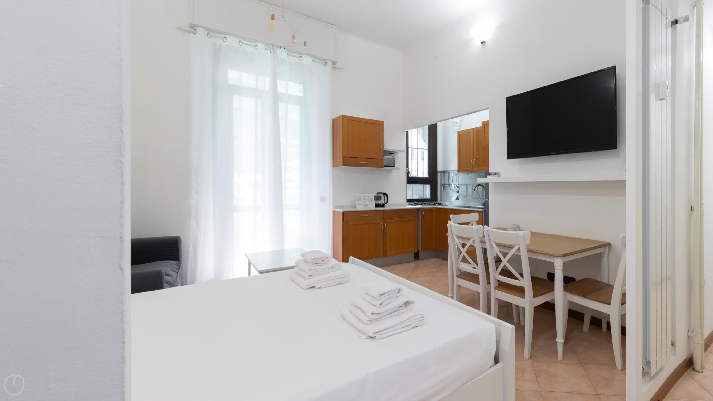 A compact hotel room with a bed, a small table with chairs, a kitchenette, and a TV mounted on the wall.