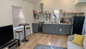 Une cuisine moderne avec des meubles gris, un four encastré et une machine à café. On y trouve un coin repas avec une table ronde et deux tabourets. Un téléviseur à écran plat est posé sur un meuble en bois. La pièce est dotée d’une grande fenêtre avec des stores et un tapis gris recouvre le sol.