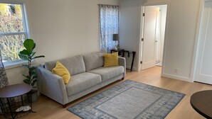 Un salon avec un canapé gris, des coussins jaunes, un tapis bleu, une plante en pot et une fenêtre donnant sur des arbres.