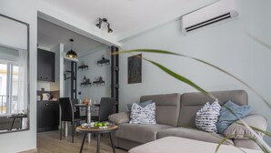 A modern living room with a grey sofa, a round table with a tray of fruit, and a kitchen in the background.