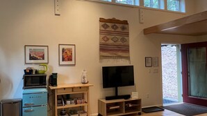 A living room with a TV, a blue refrigerator, a wooden shelf, and a window with a view of trees.
