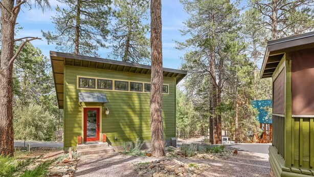 A green cabin with a red door, surrounded by trees and a gravel path.