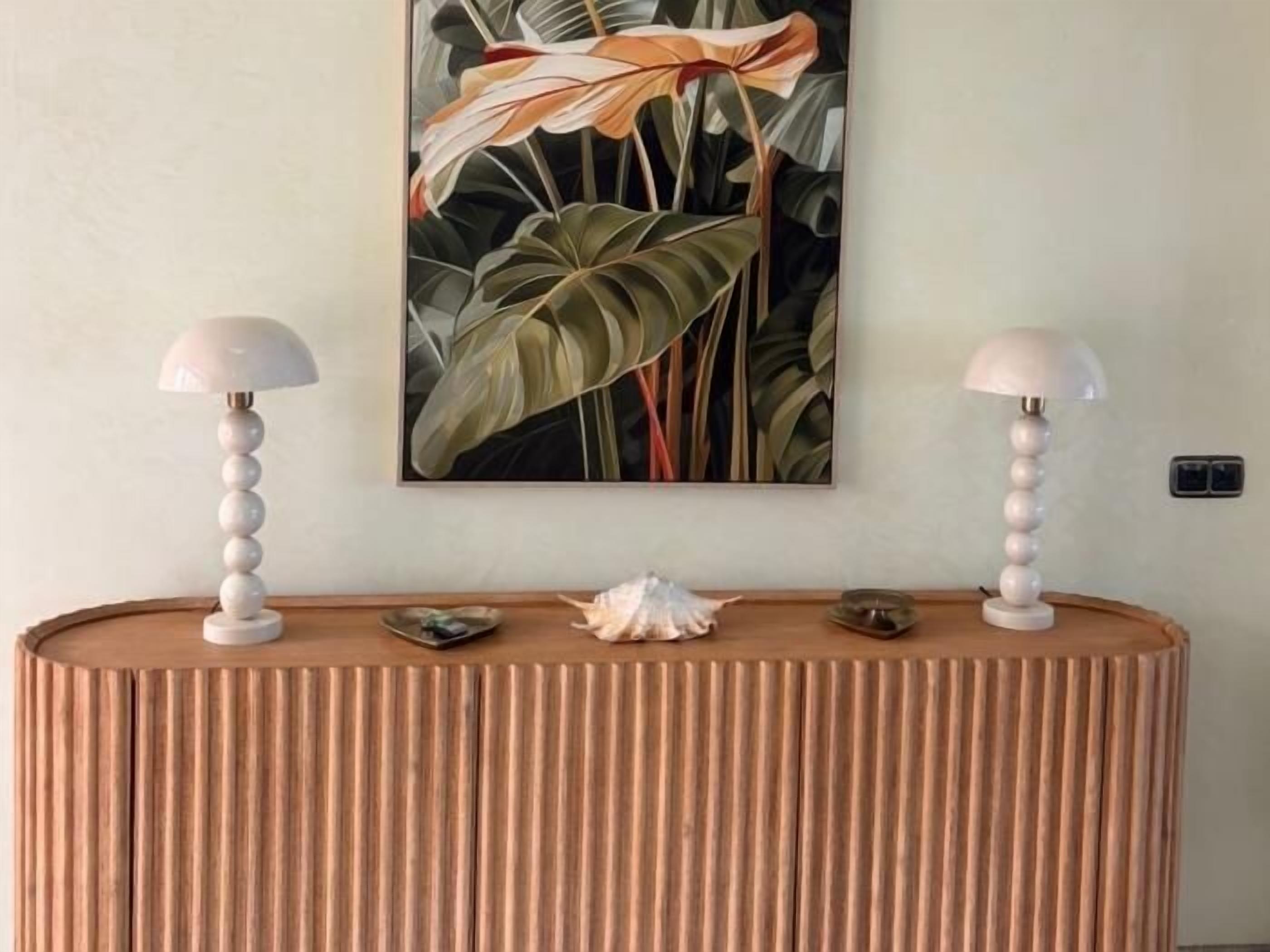 Een kamer met een ronde, ribbelige houten console tafel, twee witte lampen met kap en een groot bloemen schilderij aan de muur.