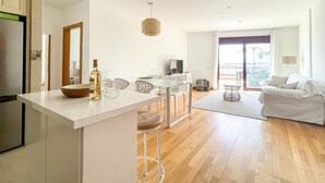 Cozinha moderna com bancada branca e banquetas, área de refeições com mesa e cadeiras, sala de estar com sofá e luminária, e varanda com vista.