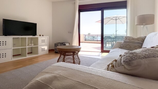 Uma sala moderna com televisão de tela plana, um sofá, uma mesa redonda pequena e uma varanda com guarda-sol.