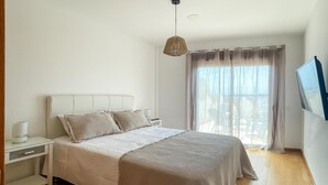 Quarto com cama, mesa de cabeceira, luminária e uma grande janela com cortinas.