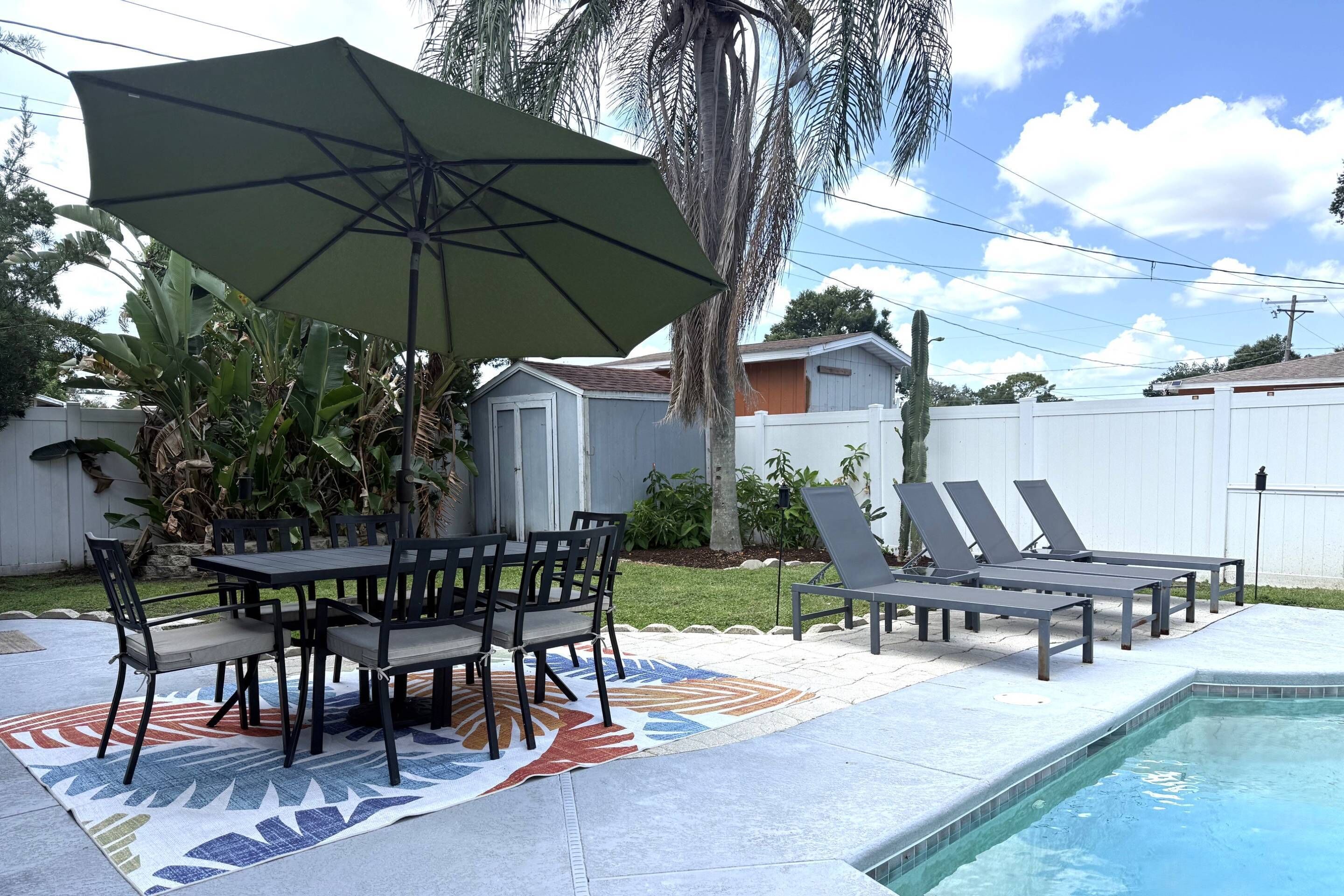 A backyard with a pool, a dining table with chairs, a lounge area with sun loungers, and a covered patio area.