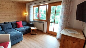 Een gezellige kamer met houten inrichting, een grijze bank met rode kussens, een klein houten tafeltje en een televisie op een houten kast.