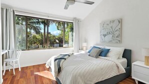 A bedroom with a bed, a ceiling fan, a large window with a view of palm trees, a white chair, and a nightstand with a lamp.