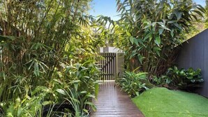 A narrow wooden pathway flanked by dense foliage and a grassy area, leading to a gated entrance.