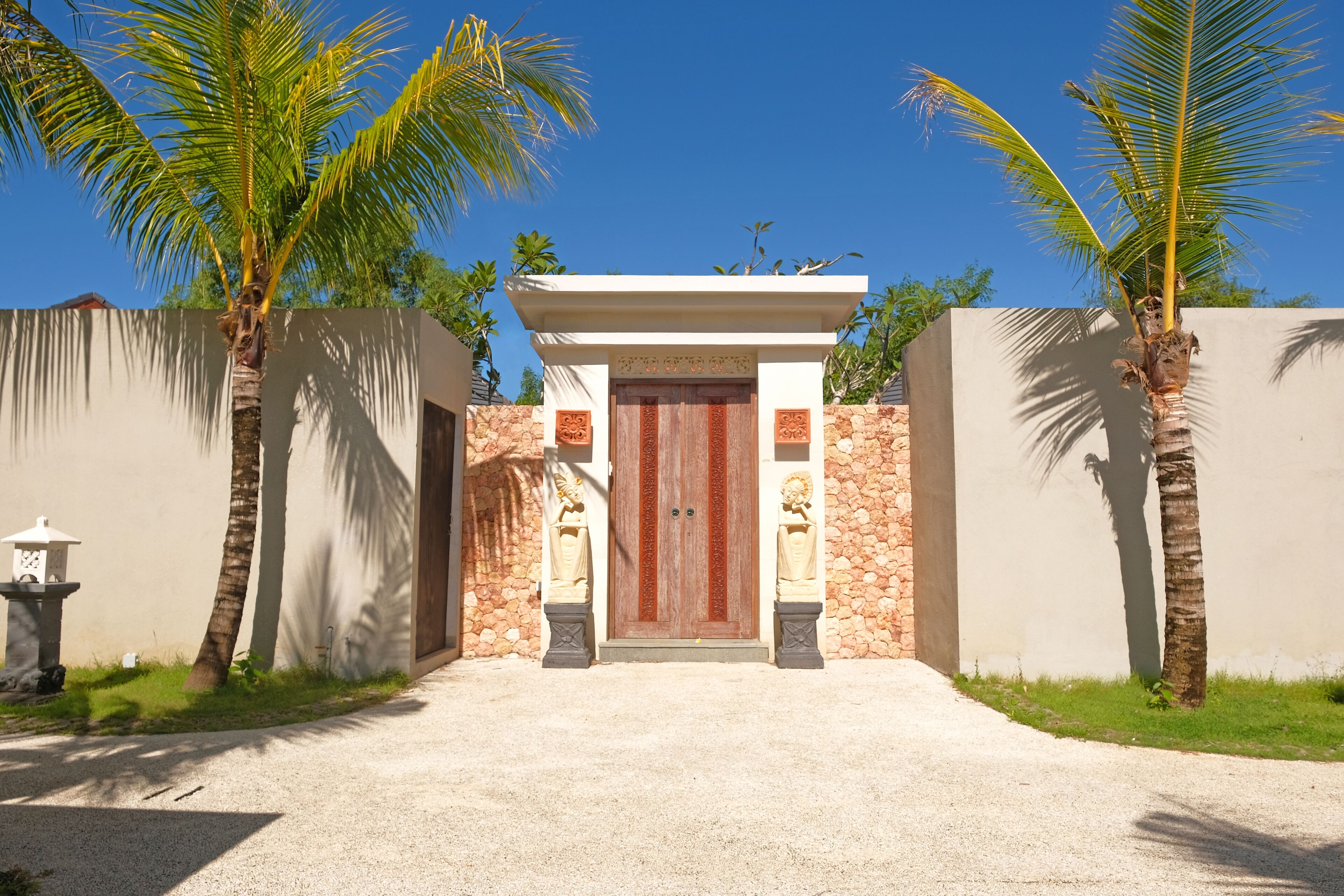 A gated entrance with a wooden door, flanked by palm trees and a stone wall.