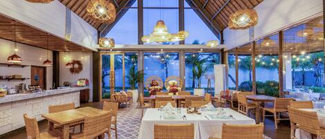 A restaurant with high ceilings, wicker furniture, and large windows overlooking a tropical landscape.
