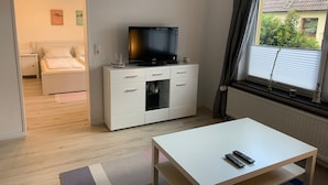 A bedroom with a bed, a TV on a white dresser, a window with curtains, and a view of greenery outside. - Ferienwohnung Zwischen Lübeck und Travemünde (Lübeck)