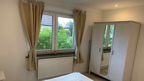 A bedroom with a bed, a wardrobe, a window with curtains, and a view of greenery outside. - Ferienwohnung Zwischen Lübeck und Travemünde (Lübeck)