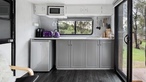 A compact kitchen with grey cabinets, a small fridge, a microwave, and a window with a view of greenery. - Glamper Lade @ The Village Green (Strath Creek)
