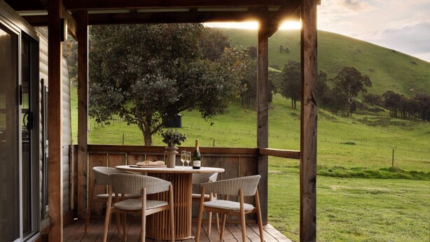A wooden deck with wicker chairs and a table, overlooking a grassy landscape with a tree and hills.