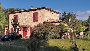 A stone building with red shutters, a car parked in front, and surrounding greenery.