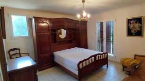 A bedroom with a wooden bed, a nightstand, a chair, a bench, a chandelier, and a window with a view of greenery.