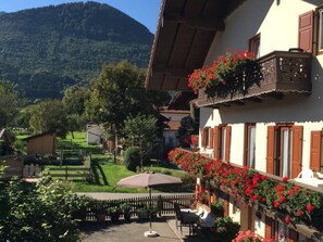 A traditional chalet with a balcony adorned with red flowers, a patio with outdoor seating, and a mountainous backdrop. - Ferienwohnung 2, 50 qm 1G (Rottau)