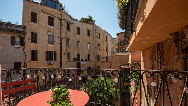 Ein Balkon mit rotem Tisch und Stühlen, einem schmiedeeisernen Geländer, und ein Gebäude mit zahlreichen Fenstern und Balkonen im Hintergrund.