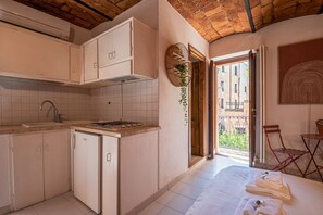 Une cuisine compacte avec des meubles blancs, un évier et une cuisinière. Il y a un balcon avec vue sur les immeubles et une porte donnant sur l’extérieur.