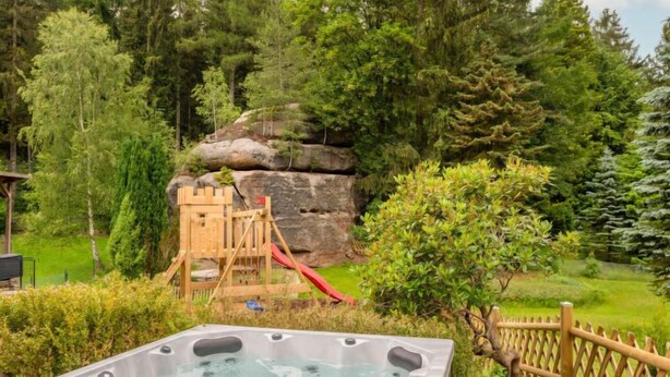A hot tub surrounded by a wooden fence, a wooden play structure with a slide, and dense greenery.