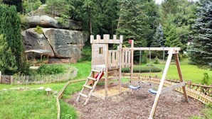 A wooden play structure with a slide and swings, surrounded by a wooden fence and a rock formation.