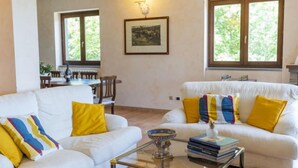Un salon avec deux canapés blancs, dont l’un est agrémenté de coussins jaunes, une table basse en verre et un tableau encadré accroché au mur.