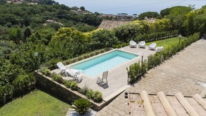 Une piscine rectangulaire entourée de chaises longues, bordée d’une terrasse pavée et d’une végétation luxuriante, et offrant une vue panoramique sur un paysage pittoresque composé de collines et d’une étendue d’eau.