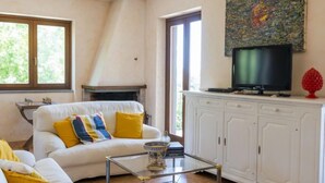 Un salon avec un canapé blanc, des coussins jaunes, une table basse en verre, une cheminée, un téléviseur posé sur un meuble et un tableau accroché au mur.