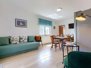 Una sala de estar moderna con un sofá verde, una mesa de comedor y sillas. Hay una ventana con cortinas, un televisor de pantalla plana montado en la pared y una lámpara de techo.