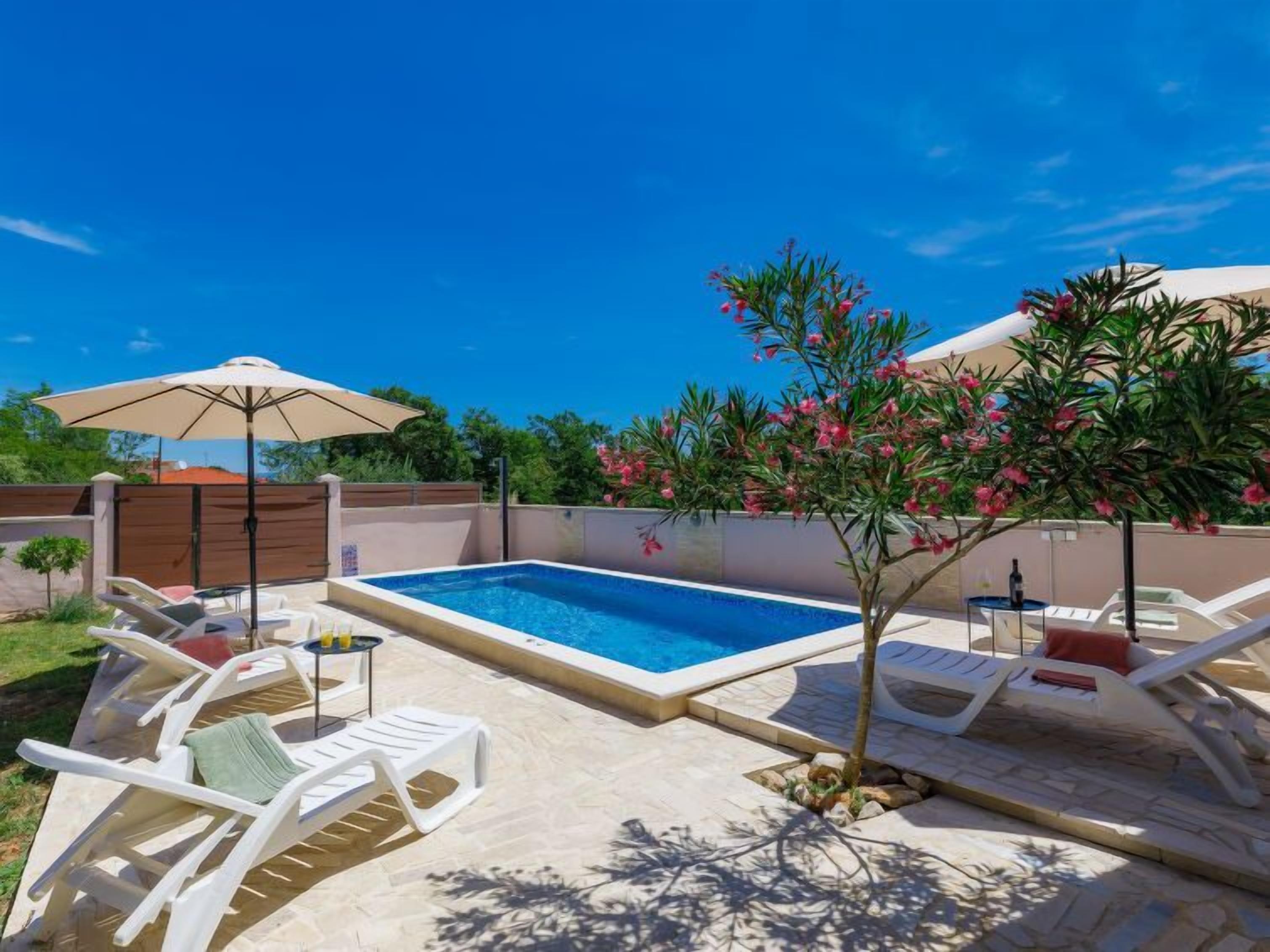 A swimming pool with lounge chairs and umbrellas, surrounded by a fence and greenery.