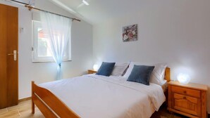A bedroom with a wooden bed, white bedding, a nightstand with a lamp, a window with sheer curtains, and a wall-mounted artwork. - Maslina by Interhome (Pula/Duga uvala)