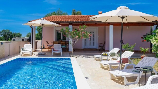 A swimming pool with lounge chairs and umbrellas, a patio area with a table and chairs, and a door leading inside. - Maslina by Interhome (Pula/Duga uvala)